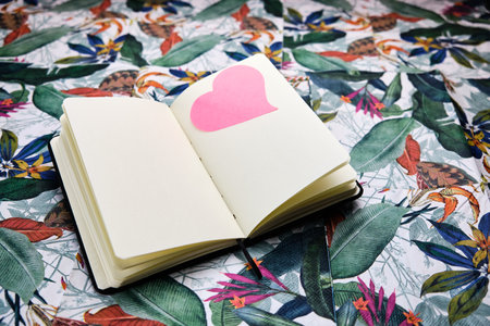 Notebook open to a blank sheet with a pink heart on a sheet, on top of a table with a tropical...
