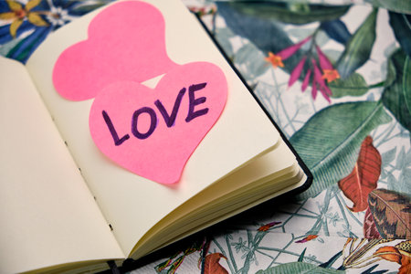 Open notebook to a blank sheet with two pink hearts on a sheet written the word Love, on top of a table with a tropical pattern.の写真素材