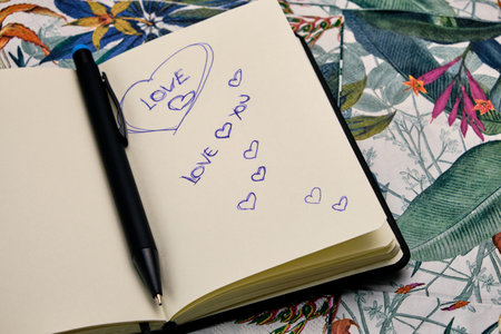 Open diary with a pen and written the word Love, on top of a table with a tropical pattern.の写真素材
