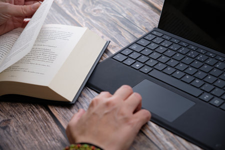 Close-up of a woman's hands studying with a laptop and a book on a wooden table.の写真素材