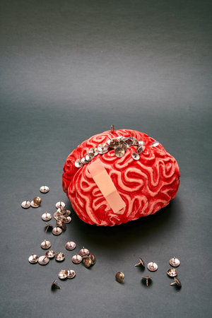 Close-up of a red rubber human brain with a bunch of thumbtacks on top of it against a dark...