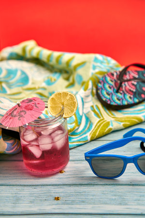 Refreshing summer drink with lots of ice and a slice of lemon, blue sunglasses, flip flops and tropical patterned beach towel on a wooden table and a red background.の写真素材
