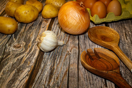 Fresh cooking ingredients are arranged on a rustic wooden table, including potatoes, onions, garlic, and eggs.の写真素材