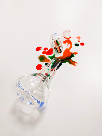 Shattered laboratory glassware with spilled colorful liquid on a white surface