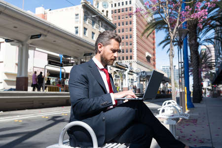 mature man businessman in suit sit on bench in outdoor city working online on laptop, freelancerの写真素材