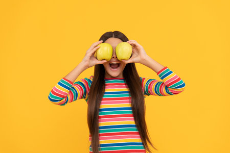 Happy girl child have fun holding apple fruit at eyes yellow background, eye healthの写真素材