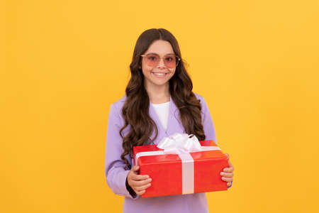 trendy cheerful kid in sunglasses and suit hold gift box on yellow background, shoppingの写真素材