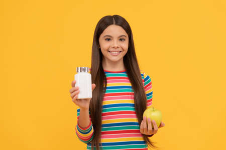 Happy girl child hold apple and supplement bottle yellow background, vitaminsの写真素材