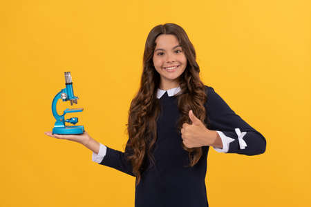 cheerful teen girl hold microscope for education on yellow background showing thumb up, chemistry.の写真素材