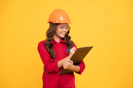 cheerful child girl in protective hard hat and glasses hold folder with paper documents, making notesの写真素材