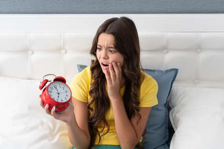 Unhappy girl child look at alarm clock sitting in bed in morning, oversleptの写真素材