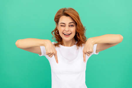winking redhead girl with curly hair pointing finger down, copy space, sales and discounts.の写真素材