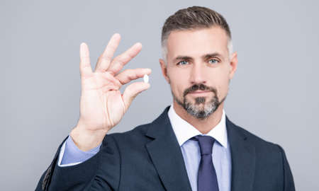 mature business man with grizzled hair in suit hold pill in hand with ok gesture, energy remedyの写真素材