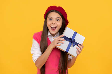 surprised teen girl in french beret hold present or gift box on yellow background, birthday