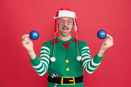 amazed man in elf costume and santa claus hat. guy in party glasses hold xmas decoration ballsの写真素材
