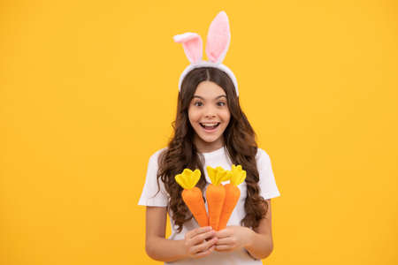 surprised child in bunny ears hold carrot on yellow background, easter