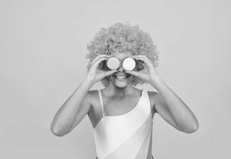 happy child in swimsuit wearing orange curly wig hair holding french macaron, sweet tooth.の写真素材