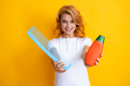 Funny portrait of redhead woman with a comb, isolated on yellow background. Woman hold bottle...