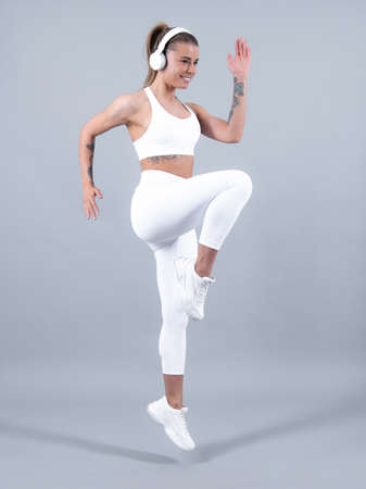 Young fitness woman jump with headphones runner stretching legs before run. Sporty woman runner on gray background. Dynamic movement. Sport music.の写真素材