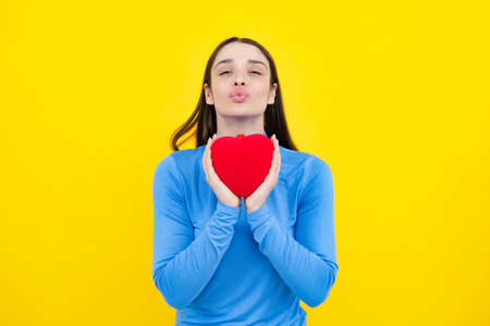 Beautiful girl holding valentines gift on yellow background. Portrait of young woman holding red paper heart. Love sign symbols for happy womens, valentines day or birthday.の写真素材