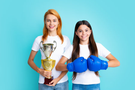 Woman power isolated blue background. Sportive young woman and her daughter wearing boxing gloves, holding winning prize, showing trophy, emotional excited and happy.の写真素材