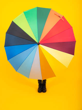 Autumn kids concept. Child teenager girl covered under rainbow umbrella in autumn black leather boots on yellow studio background. Autumn kids trendy shoes.の写真素材