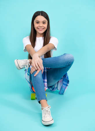 Fashion teen girl wearing trendy t-shirt and jeans sit on skateboard, posing on blue background....