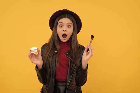 shocked kid in hat hold makeup brush and powder on yellow background, skincare