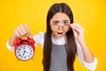 Teen student girl hold clock isolated on yellow background. Time to school. Teenager child with alarm clock showing time, late awakening. Surprised teenager girl.の写真素材