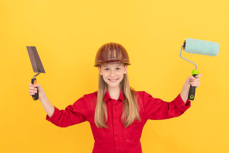 happy teen child in builder hard hat with paint roller and spatula on yellow wallの写真素材