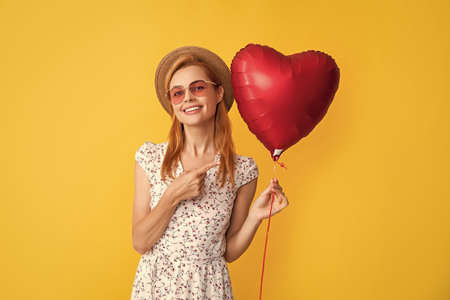 happy young woman in straw hat and sunglasses point finger love heart balloon on yellow backgroundの写真素材