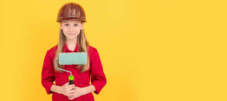 smiling teen child in builder hard hat with paint roller on yellow wall. Child builder in helmet...