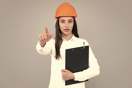 Architect woman in helmet pointing on copy space. Young woman in helmet hold clipboard on gray...