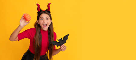 amazed kid with pumpkin and bat wearing devil horns on yellow background. Halloween kid girl portrait, horizontal poster. Banner header with copy space.の写真素材