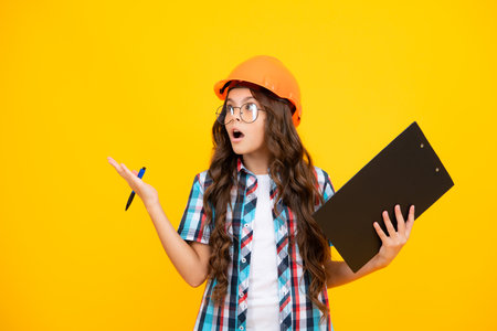 Shocked surprised teenager girl. Teenager child painter in helmet with clipboard. Child in hard hat. Kid engineer on construction site. Renovation and repair.の写真素材