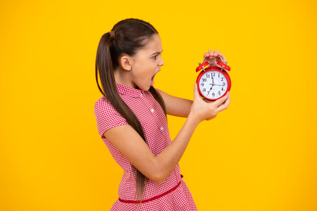 Unhappy sad teenager girl. Child student girl with clock isolated on yellow background. Child back to school. Education and time concept.の写真素材