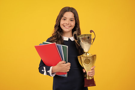 Schoolgirl in school uniform celebrating victory with trophy. Teen holding winning award prize on yellow background. Child hold books with gold trophy or winning cup. Education graduation, victory.の写真素材