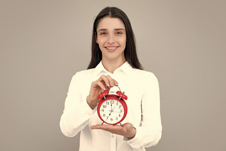 Woman holds a clock. Emotional young female face. Portrait of young surprised woman with alarm clock on grey background.の写真素材