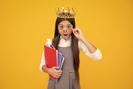 Surprised teenager girl. Schoolgirl in school uniform and crown celebrating victory on yellow background. School child hold books. Education graduation, victory and success.の写真素材