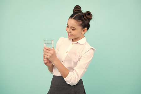 Cute teen girl drinking water from glass on blue background. Daily life health. Drink water for health care and body balance. Happy teenager, positive and smiling emotions of teen girl.の写真素材