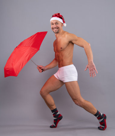 Crazy santa with umbrella. Naked gay santa in santa head funny posing on gray studio background. Muscular body, male torso.の写真素材