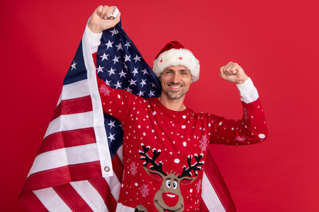 Santa hold american flag. Xmas in united states, usa. Portrait of middle aged man in sweater isolated over red background. Concept of holidays, happiness, emotions and Christmas celebration.の写真素材