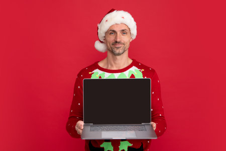 Man in Christmas sweater and hat on color background. Santa with laptop. Middle aged santa hold laptop with blank screen.の写真素材