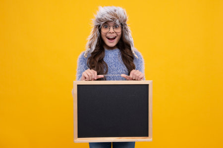 Teenager girl hold in warm winter hat hold blackboard chalkboard with copy space on yellow background. Excited face, cheerful emotions of teenager girl.の写真素材