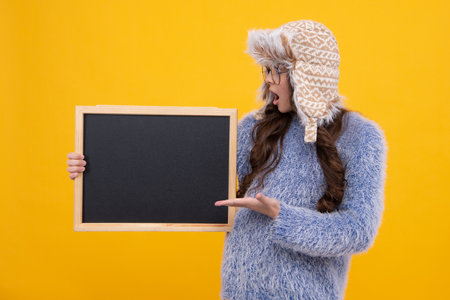 Teenage girl child in warm winter hat and sweater holding blackboard, isolated on a yellow background. Winter school sale, copy space, mockup. Surprised face, surprise emotions.の写真素材