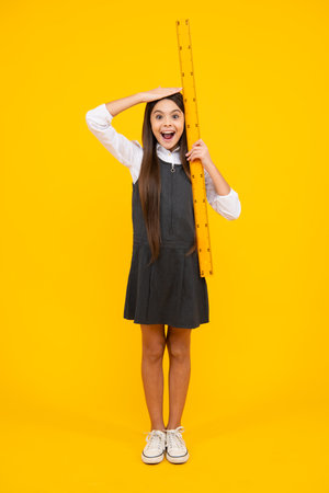 Kid height measure, growth measurement. Schoolgirl holding measure for geometry lesson, isolated on yellow background. Student study math. Excited face, cheerful emotions of teenager girl.の写真素材