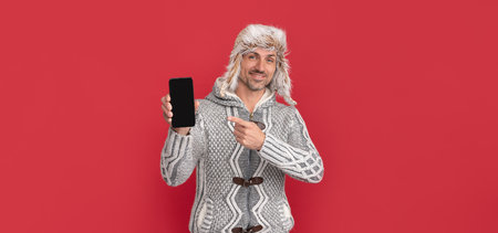 happy grizzled guy in sweater and earflap hat pointing finger on smartphone, presenting product.