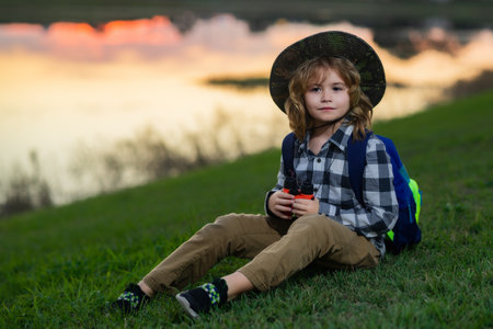 Cute blond kid with binoculars wearing explorer hat and backpack on nature. Child explorer hiking and adventure.の写真素材