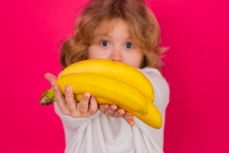 Child hold banana in studio. Studio portrait of cute kid boy with bananas isolated on red background.の写真素材