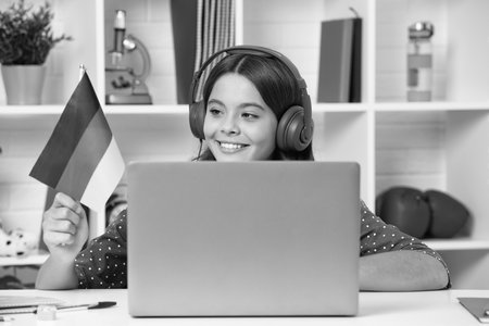 A young school girl student sitting at the table, using laptop when studying. Happy girl hold german flag, positive and smiling emotions.の写真素材
